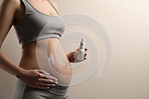 Pregnant woman applying cosmetic product on belly against beige background, closeup. Space for text
