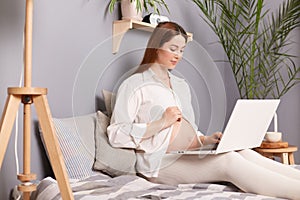 Pregnant woman with bare belly sitting on bed using laptop future mom expecting child working on computer answering email messages