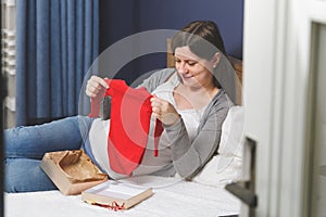 A pregnant woman in bed opens presents from a baby shower, holding baby clothes in her hands.