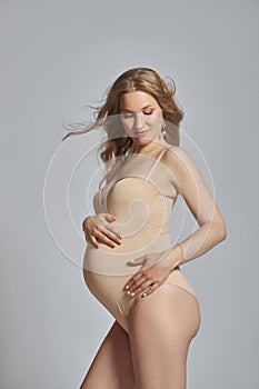 Pregnant woman in a beige bodysuit smiles, cradling her belly with both hands while standing indoors against a gray background photo