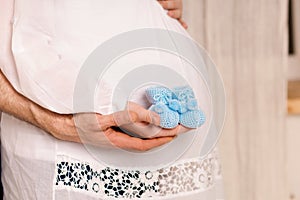 Pregnant woman with belly, man hugging big tummy, holding knitted shoes for the baby