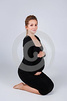 A pregnant woman in a black dress is sitting on the floor in the studio on a gray background. Expectant mother is expecting a baby