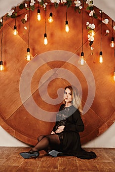 Pregnant woman in black dress touching her belly on decoration wall with retro light bulbs. Motherhood, pregnant, people and