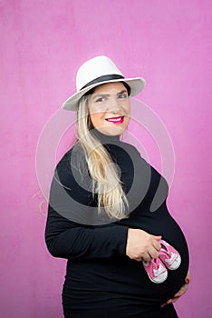 a pregnant woman in a black outfit and hat holding pink baby shoes