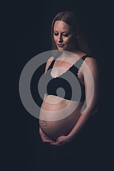 Pregnant woman in black underwear holding belly on black background