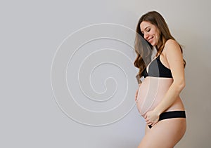 Pregnant woman in black underwear holding her belly in grey copy space background.