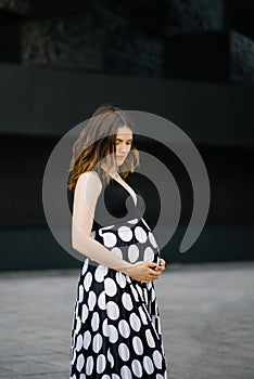 A pregnant woman in a black and white dress holds her hands on her pregnant belly