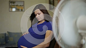 A pregnant woman in a blue dress is sitting in front of a white fan.