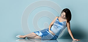 Pregnant woman in blue silk dress or nightie. She smiling, put hand on her belly, posing sitting on floor of blue photo background