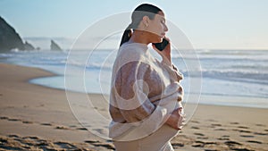 Pregnant woman calling cellphone at beach caressing big belly. Girl talking