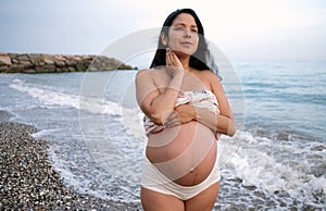 Pregnant woman caressing her bare belly with her hands standing on a beach at sunset with a delicate evening light glowing on the