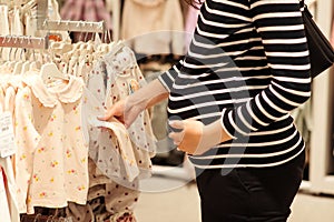 Pregnant woman choosing dress and bodysuits at clothing store. Baby fashion, shopping time, sale and pregnancy concept