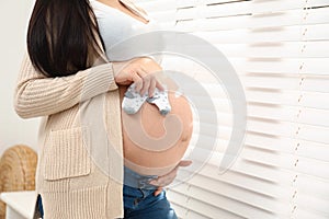 Pregnant woman with cute baby socks near window at home