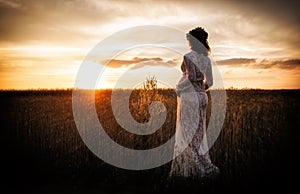 A pregnant woman in a delicate negligee in a field enjoys a beautiful sunset. Beauty and tender motherhood