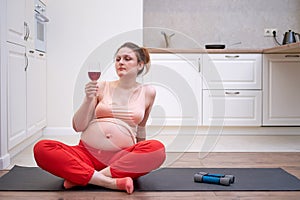 Pregnant woman doing fitness at home and looking at red wine in a glass