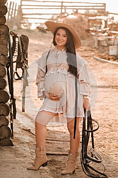 a pregnant woman in a dress and hat in the countryside holds a horse bridle in her hand