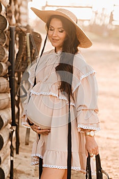 a pregnant woman in a dress and hat in the countryside holds a horse bridle in her hand