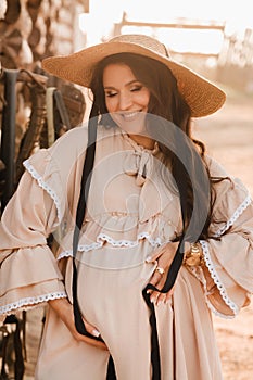 a pregnant woman in a dress and hat in the countryside holds a horse bridle in her hand