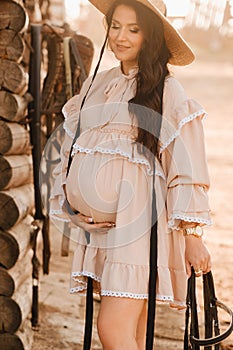 a pregnant woman in a dress and hat in the countryside holds a horse bridle in her hand