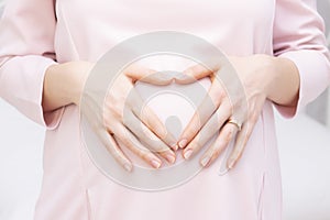 A pregnant woman in a dress holds her hands on her stomach in the shape of a heart. The concept of pregnancy