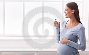 Pregnant Woman Drinking Glass Of Water Standing Against Window Indoor