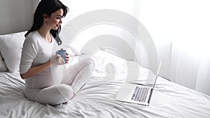 Pregnant Woman Drinking Tea, Using Computer At Home In Bedroom