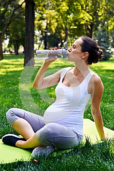 Pregnant woman drinking water on yoga mat in park