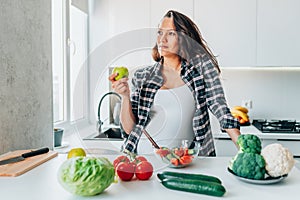 Pregnant woman eating apple while preparing vegetable salad in kitchen at home