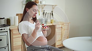 Pregnant woman eating dessert and smiling while sitting at table in home room spbd.