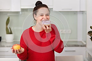 Pregnant woman eating sweet cake and holding apple in another hand, stands wearing red casual sweater in front white kitchen