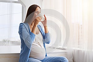 Pregnant woman enjoying cup of tea while sitting next to the win