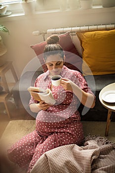Pregnant woman enjoying at home reading book and holding cup of coffee.