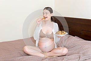 Pregnant woman enjoys eating cookies resting in bed at home. Unhealthy sweet pastry during pregnancy concept