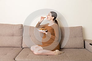 Pregnant woman enjoys eating cookies resting on the sofa at home. Unhealthy sweet pastry during pregnancy concept