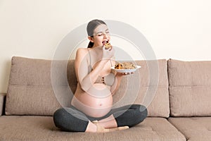 Pregnant woman enjoys eating cookies resting on the sofa at home. Unhealthy sweet pastry during pregnancy concept