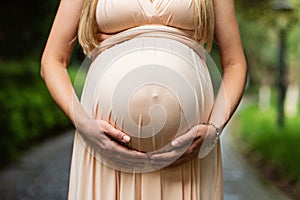 Pregnant woman in evening dress holds hands on belly outdoor in summer park. Pregnancy, maternity, preparation and