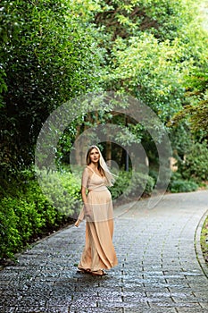 Pregnant woman in evening dress standing outdoor in summer park. Pregnancy, maternity, preparation and expectation