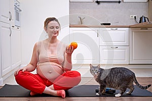 A pregnant woman exercises at home and holds a fruit in her hand as a symbol of healthy food during pregnancy