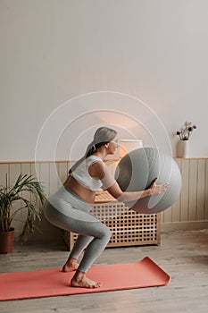 Pregnant woman exercising with fit ball at home, making squats with fitball.