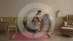 Pregnant woman exercising with fit ball at home, making squats with fitball.