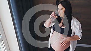 A pregnant woman, a expectant mother, stands near the window and drinks water. Health and pregnancy.