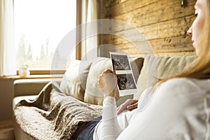 Pregnant woman expecting newborn and holding ultrasound while lying comfortably on couch