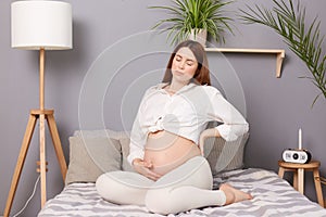 Pregnant woman experiencing lower back pain causing discomfort while she sitting on her bed at home, possibly indicating the onset