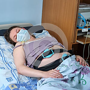 A pregnant woman in face mask gives birth in a hospital with a drip and a cardiotocograph machine. A woman on a clinic bed during