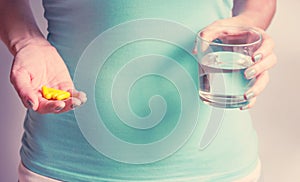 A pregnant woman first trimester in a blue T-shirt holds several medicine capsules, and in the second a glass of water. IVF.