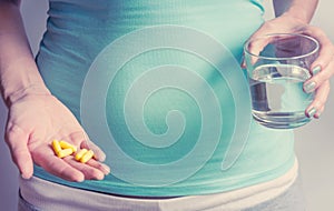A pregnant woman first trimester in a blue T-shirt holds several medicine capsules, and in the second a glass of water. IVF.