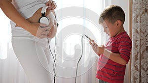 Pregnant woman gives music to baby in belly through headphones, mobile phone into hands of little boy standing beside