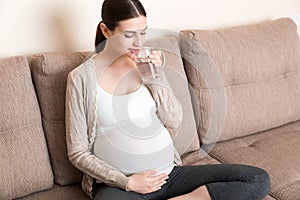 Pregnant woman with a glass of water sitting on sofa at the home