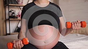 Pregnant woman goes in for sports with dumbbells sitting at home on the bed.