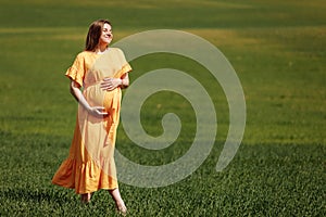 Pregnant woman among the green field. Romantic pregnant woman outside, in the field and among greenery like a fairytale